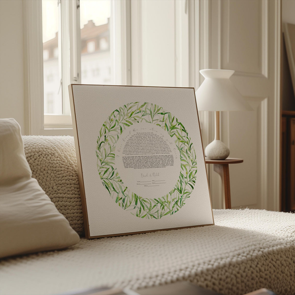 A framed watercolor painting of a wreath with green leaves is displayed on a white couch in a room with a lamp and window in the background.